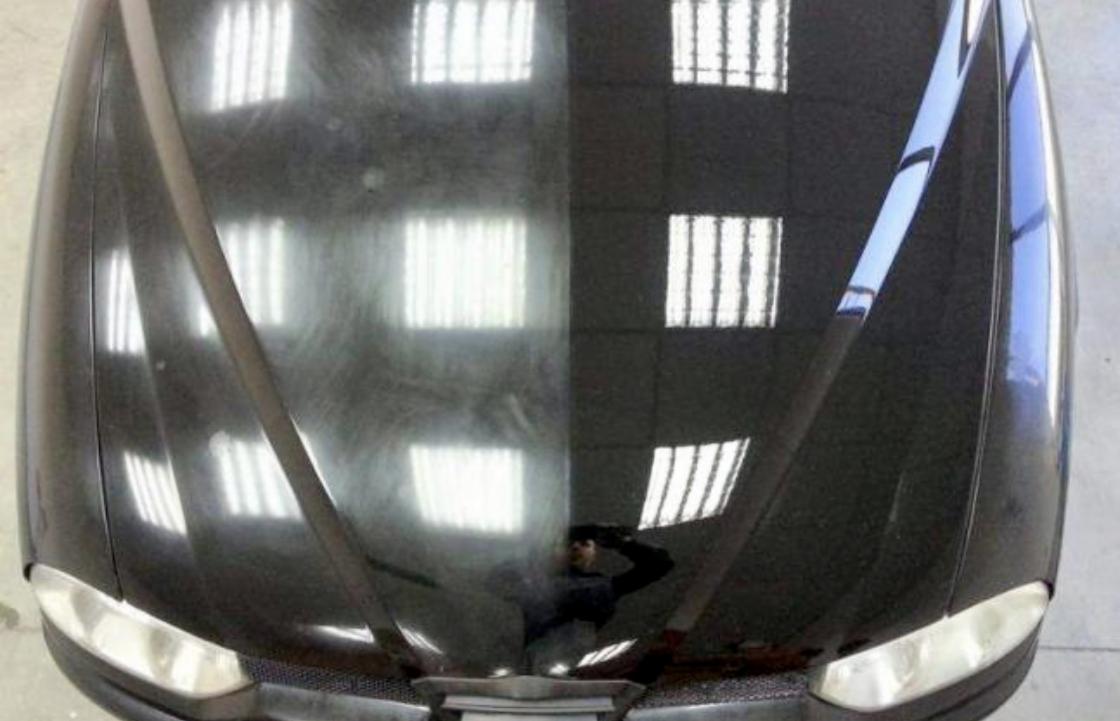 Częściowo wypolerowana maska czarnego samochodu, widoczna różnica w połysku i usunięciu zarysowań, odbicie okien w lakierze.