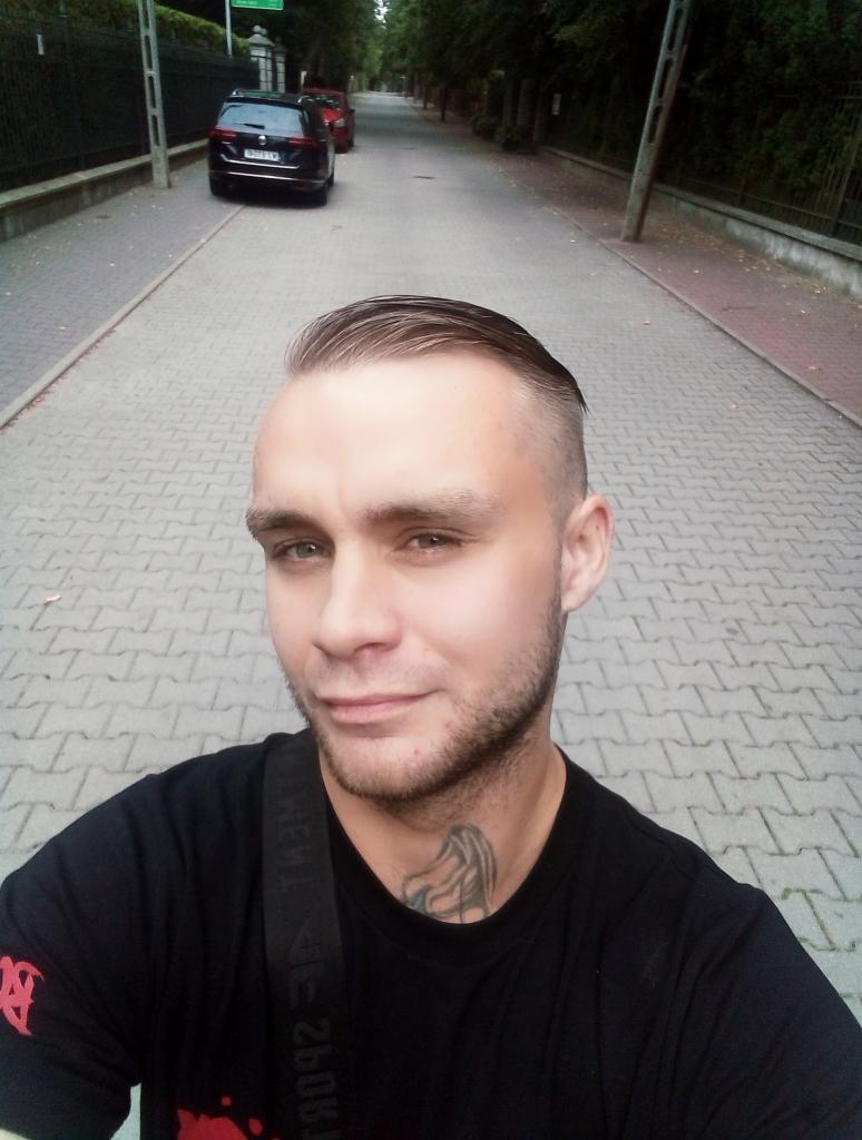 Mężczyzna z tatuażem na szyi robi selfie na tle brukowanej ulicy z zaparkowanymi autami i zielenią w tle. Ujęcie z dołu.