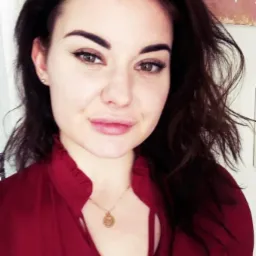 Portret kobiety w bordowej koszuli z delikatnym makijażem i złotym naszyjnikiem, brązowe włosy opadają na ramiona, subtelny uśmiech.