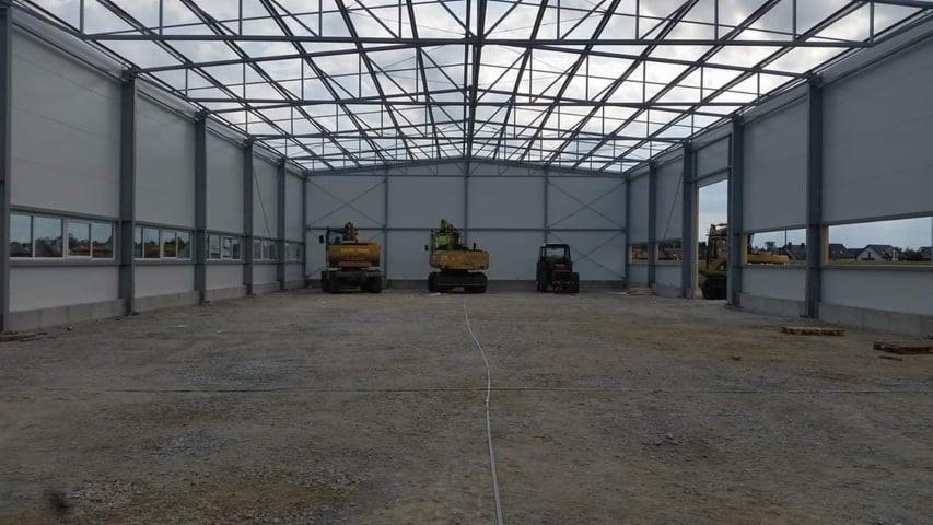 Wnętrze hali w trakcie budowy, z widocznymi trzema maszynami budowlanymi: dwiema koparkami i małym traktorem, na nierównym, żwirowym podłożu. Konstrukcja stalowa dachu jest odsłonięta, a ściany...
