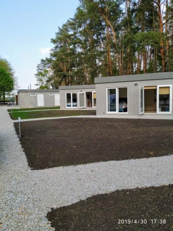 Przygotowany teren pod budowę modułowych domków letniskowych z wysypanym szarym żwirem na ścieżce, otoczony lasem iglastym i błękitnym niebem.