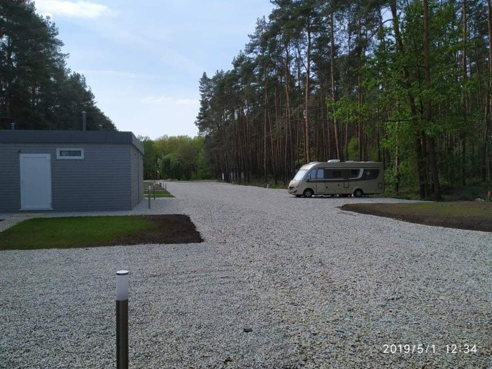 Ukończony plac postojowy z szarego grysu otoczony lasem, z widocznym budynkiem gospodarczym i zaparkowanym kamperem.