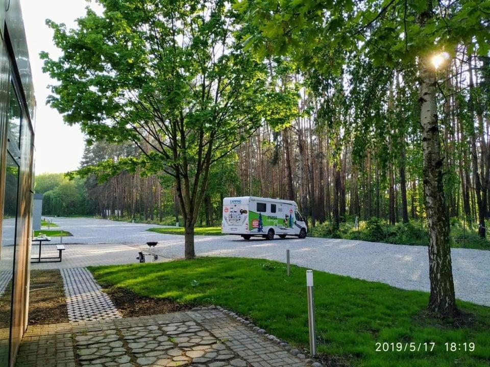 Kemping z utwardzoną, żwirową drogą dojazdową, otoczony drzewami i zielenią, z widocznym kamperem w tle.