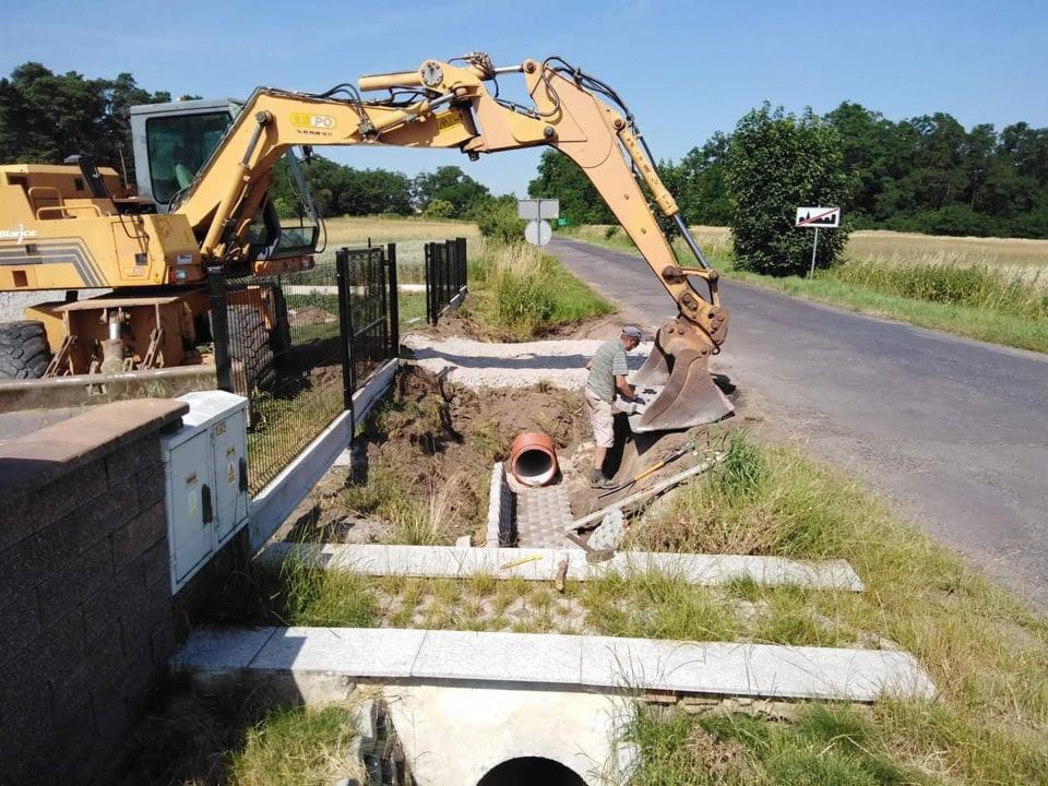Żółta koparka przy drodze, pracownik montuje rurę w wykopie, widoczne elementy betonowe i ogrodzenie, w tle pole i drzewa.