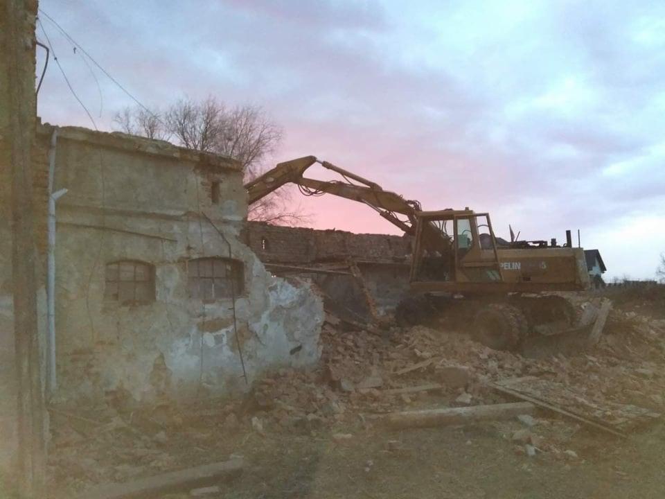 Żółta koparka gąsienicowa na gruzowisku podczas rozbiórki starego budynku, z fragmentami ścian i okien, w tle niebo o zmierzchu z różowymi odcieniami.