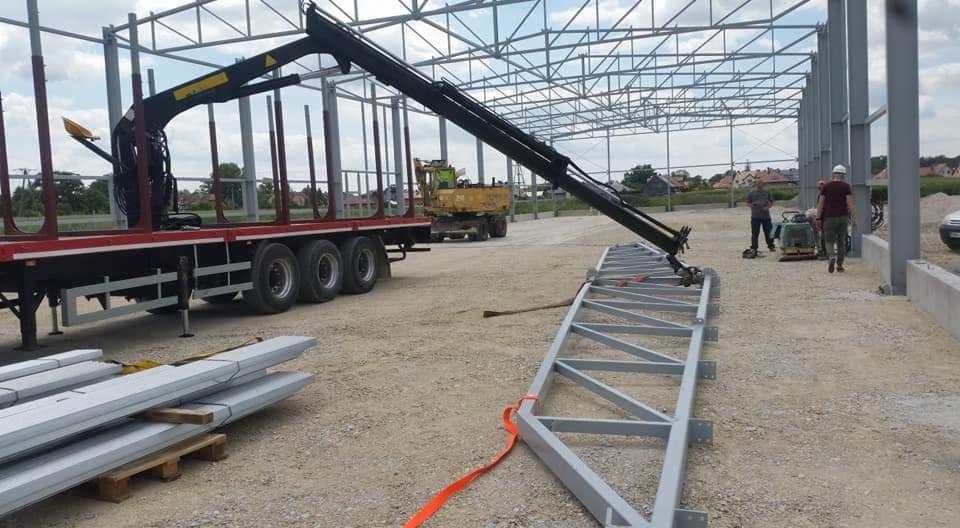 Montaż stalowej konstrukcji hali: ciężarówka z dźwigiem przygotowuje elementy, w tle widoczna koparka i pracownicy z zagęszczarką.