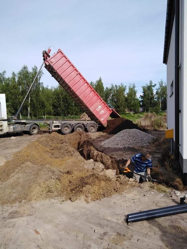 Wyładunek ziemi z czerwonej wywrotki obok wykopu, pracownik wyrównuje dno łopatą, widoczne rury kanalizacyjne i hałda żwiru.