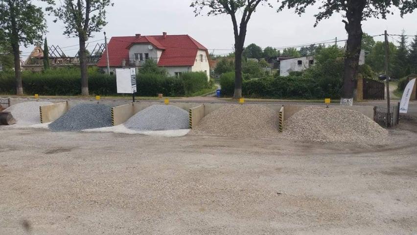 Skladowisko różnych rodzajów kruszyw budowlanych: piasek, żwir, kamień w oddzielnych boksach z betonowymi ściankami, na tle zabudowań mieszkalnych i drzew.
