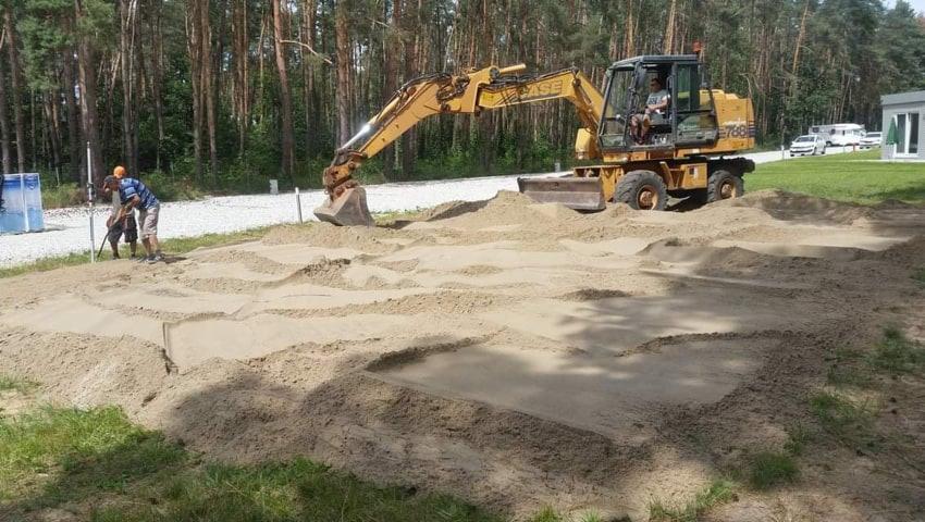 Żółta koparka CASE 768B profiluje teren z piasku, w tle dwóch mężczyzn z niwelatorem optycznym, widoczny las i zabudowania.