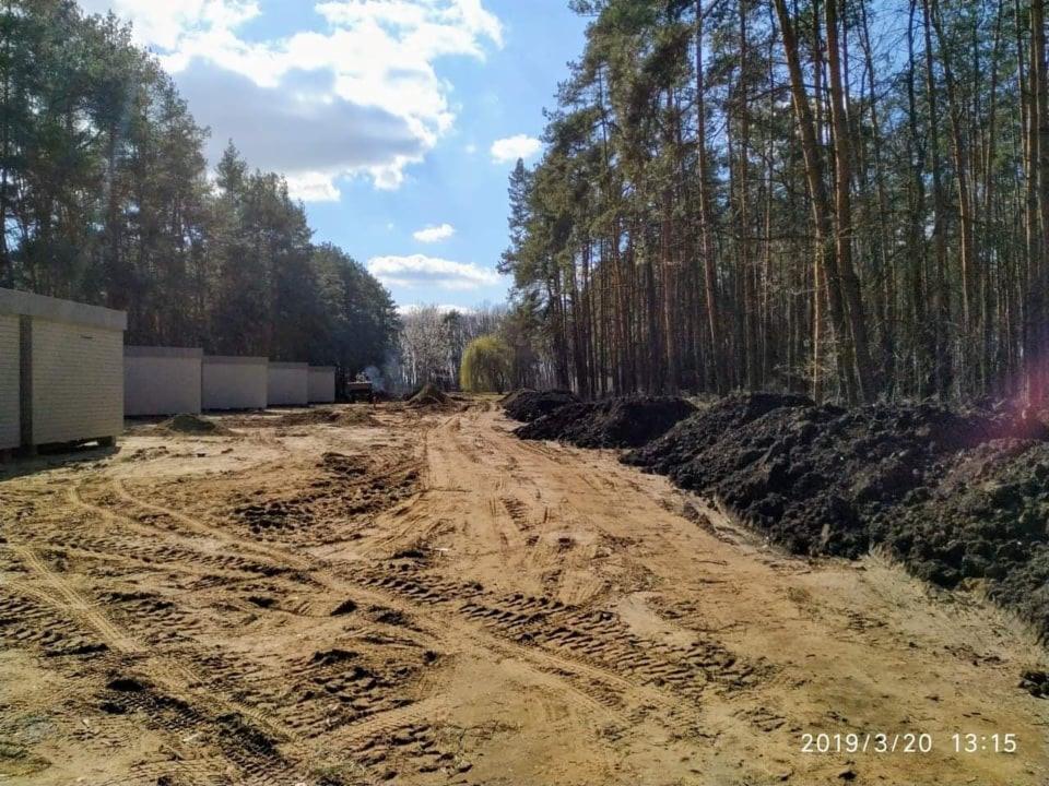 Teren przygotowany pod budowę w lesie, z widocznymi śladami opon maszyn budowlanych, hałdami ziemi i ustawionymi tymczasowymi budynkami gospodarczymi.