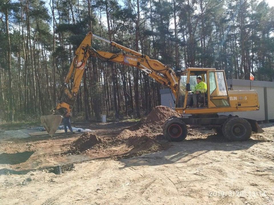 Żółta koparka kołowa marki ELIN z operatorem w jaskrawej kamizelce wykonuje prace ziemne na piaszczystym terenie w otoczeniu wysokich sosen, widoczny wykop i pryzma ziemi.