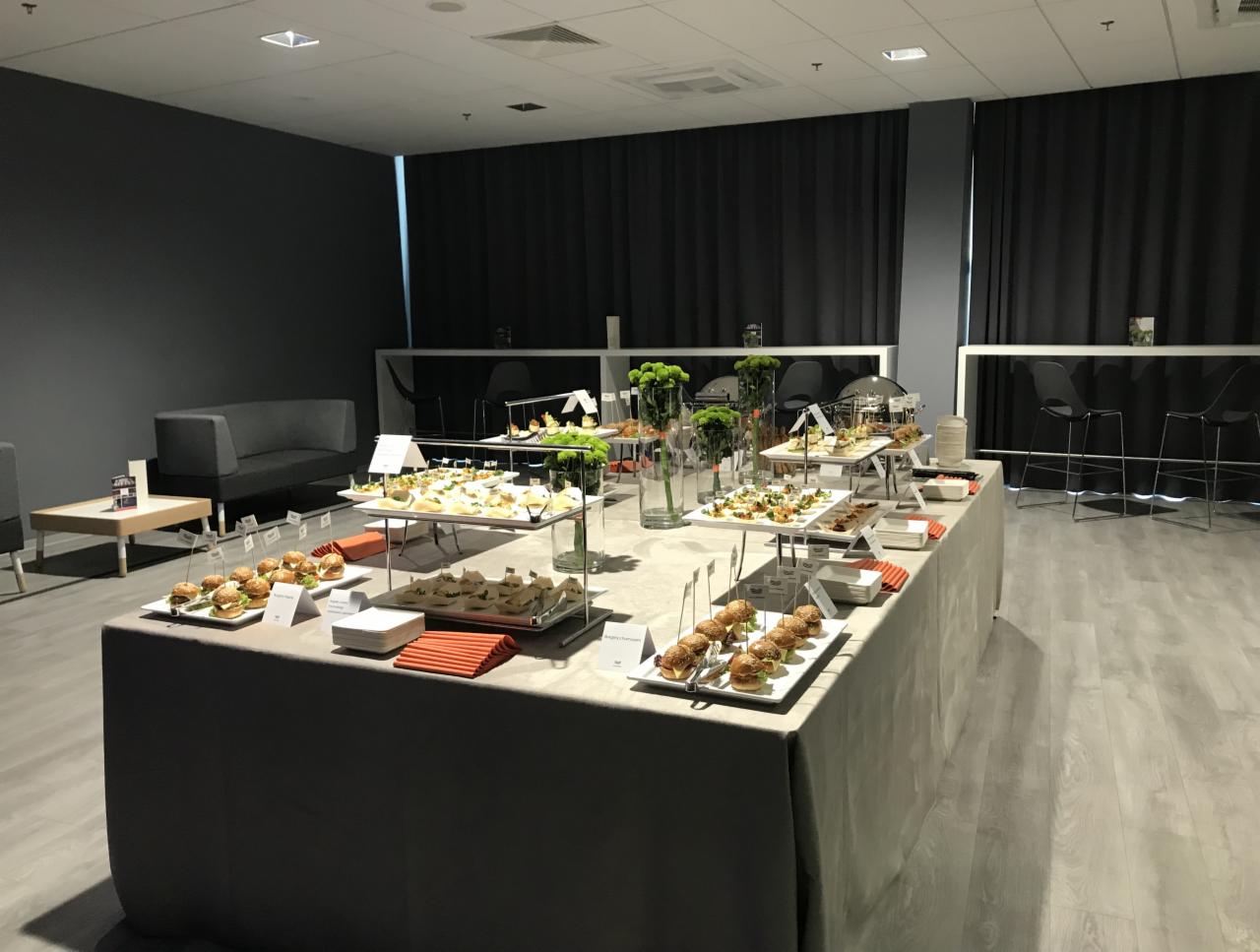 Stół cateringowy z mini burgerami, kanapeczkami i dekoracjami kwiatowymi w nowoczesnym wnętrzu z szarą ścianą i zasłonami.