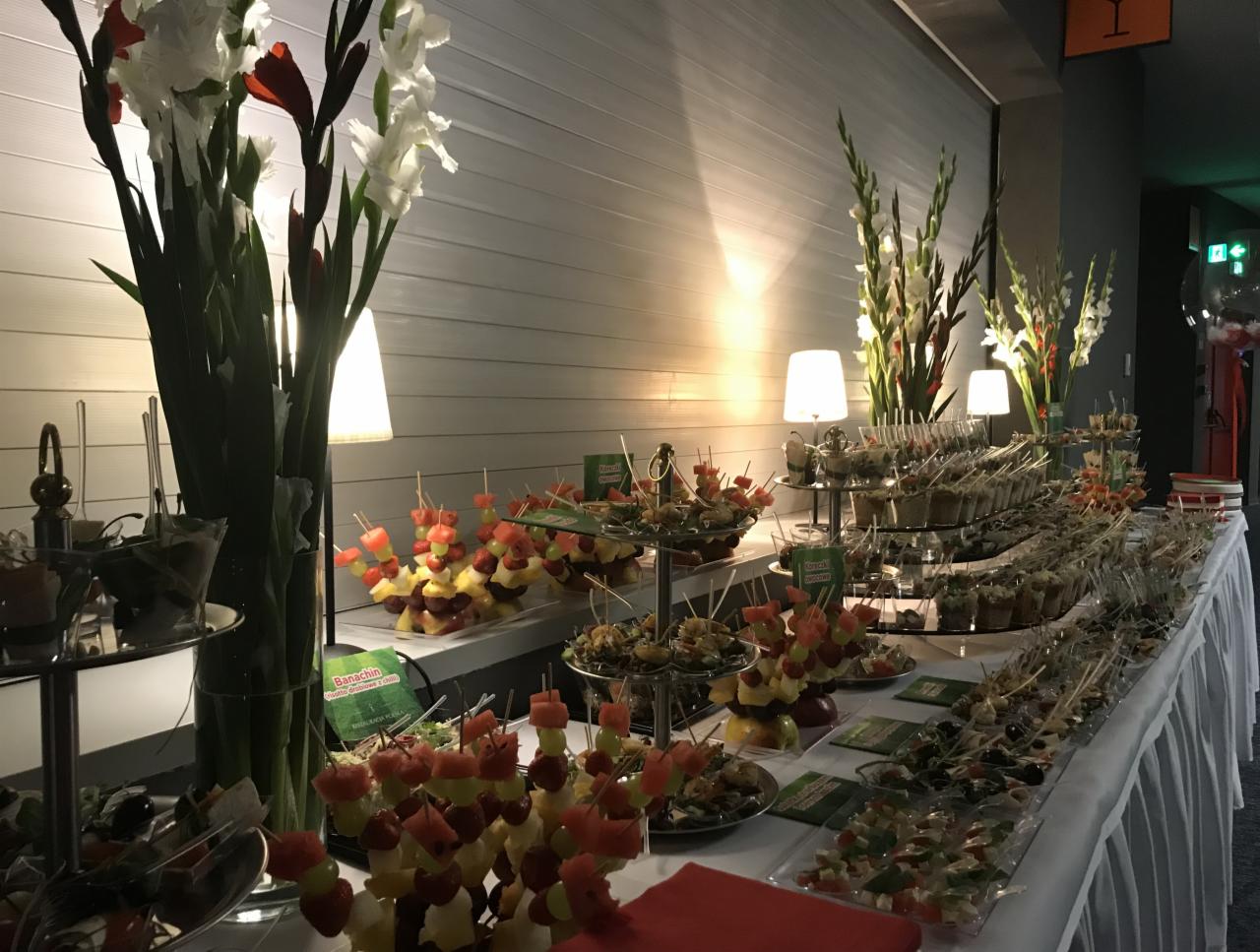 Stół cateringowy z przekąskami, owocowymi szaszłykami i kwiatami w wazonach, oświetlony lampkami, z widocznym logo firmy cateringowej na małych tabliczkach.