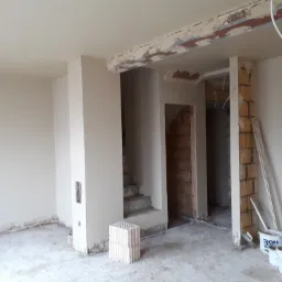 Wnętrze w Damasławku w trakcie prac wykończeniowych: ściany pokryte jasną warstwą, widoczne fragmenty surowej cegły, dwa wiadra z zaprawą, listwy przypodłogowe i przewody wystające z sufitu.