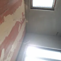 Wnętrze w trakcie prac wykończeniowych: ściana z fragmentami starej farby i nałożoną warstwą wyrównującą, sufit przygotowany pod malowanie, widoczne okno dachowe i przewody elektryczne.