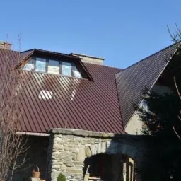 Dom z brązowym, metalowym dachem z oknem dachowym, kamiennym wejściem i fragmentem osoby w lewym dolnym rogu. Widok z dołu na błękitne niebo.