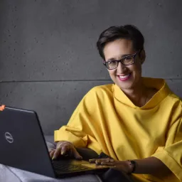 Uśmiechnięta kobieta w okularach i żółtej bluzce siedzi na szarej sofie z laptopem Dell na kolanach, pracując i patrząc w obiektyw.