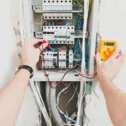 Elektryk mierzy napięcie w skrzynce rozdzielczej za pomocą multimetru, widoczne kable i zabezpieczenia