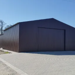 Gotowa hala z brązowej blachy trapezowej z bramą wjazdową, widok z zewnątrz na tle błękitnego nieba i wiejskich zabudowań.