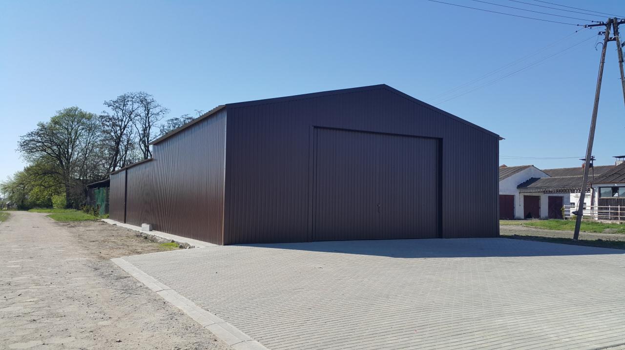 Gotowa hala z brązowej blachy trapezowej z bramą wjazdową, widok z zewnątrz na tle błękitnego nieba i wiejskich zabudowań.