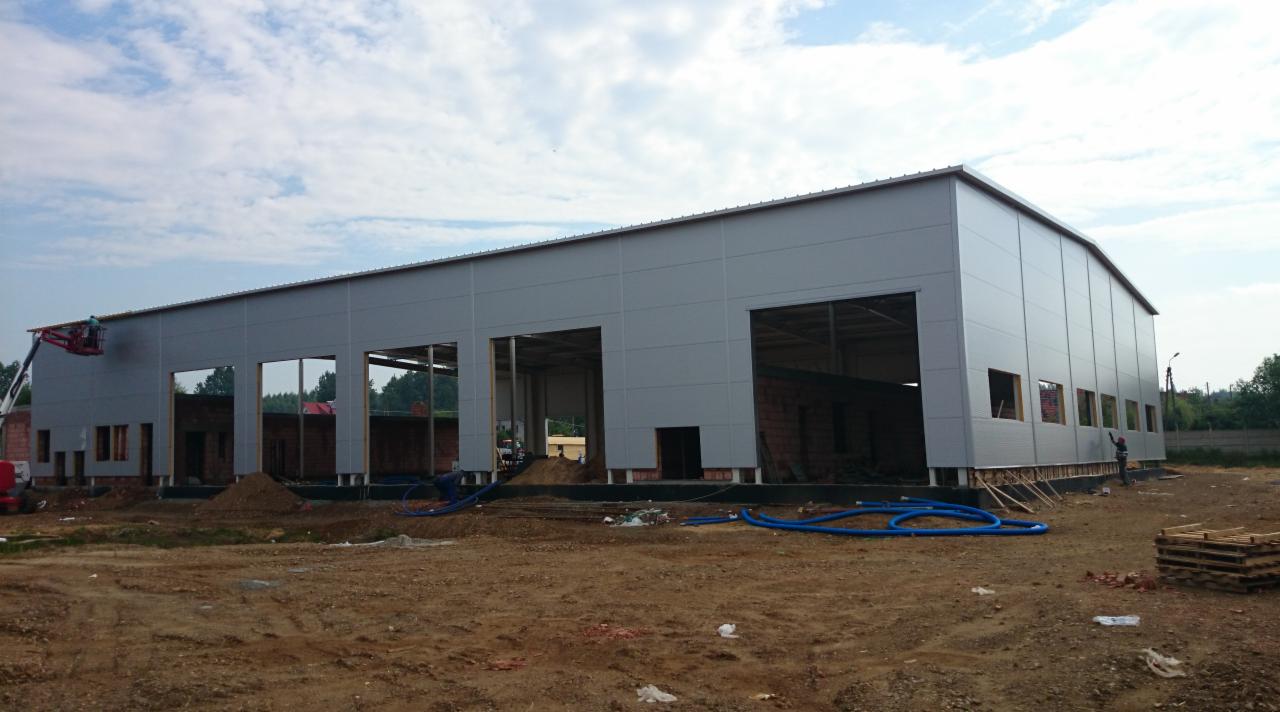 Budowa hali stalowej z płyt warstwowych w trakcie montażu, widoczna podnośnik koszowy i częściowo wzniesione ściany z cegły