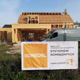 Konstrukcja domu w technologii szkieletowej w trakcie budowy, widoczny szkielet drewniany, dach w budowie, na pierwszym planie baner reklamowy systemów kominowych Schiedel oraz zaparkowany szary bus.