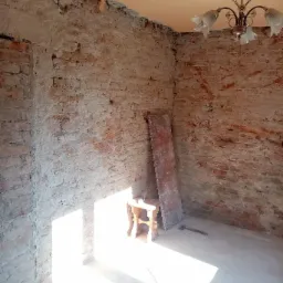 Wnętrze remontowanego pokoju z odsłoniętą cegłą, drewnianym stołkiem, deską i siekierą leżącą na podłodze, oświetlone jasnym światłem wpadającym przez okno.