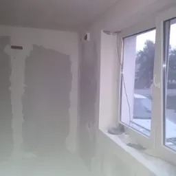 Wnętrze w trakcie prac wykończeniowych: ściany po szpachlowaniu, widoczne przewody elektryczne przy oknie z widokiem na ulicę i zaparkowany samochód.