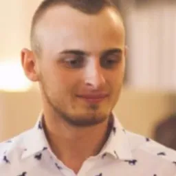 Portret młodego mężczyzny w koszuli z wzorem, z krótką fryzurą, patrzącego w dół, w neutralnym oświetleniu.