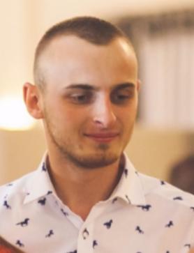 Portret młodego mężczyzny w koszuli z wzorem, z krótką fryzurą, patrzącego w dół, w neutralnym oświetleniu.