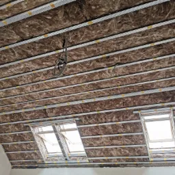 Poddasze z widoczną izolacją z wełny mineralnej między metalowymi profilami, zabezpieczonymi żółtą taśmą, oraz trzema oknami dachowymi w trakcie montażu.