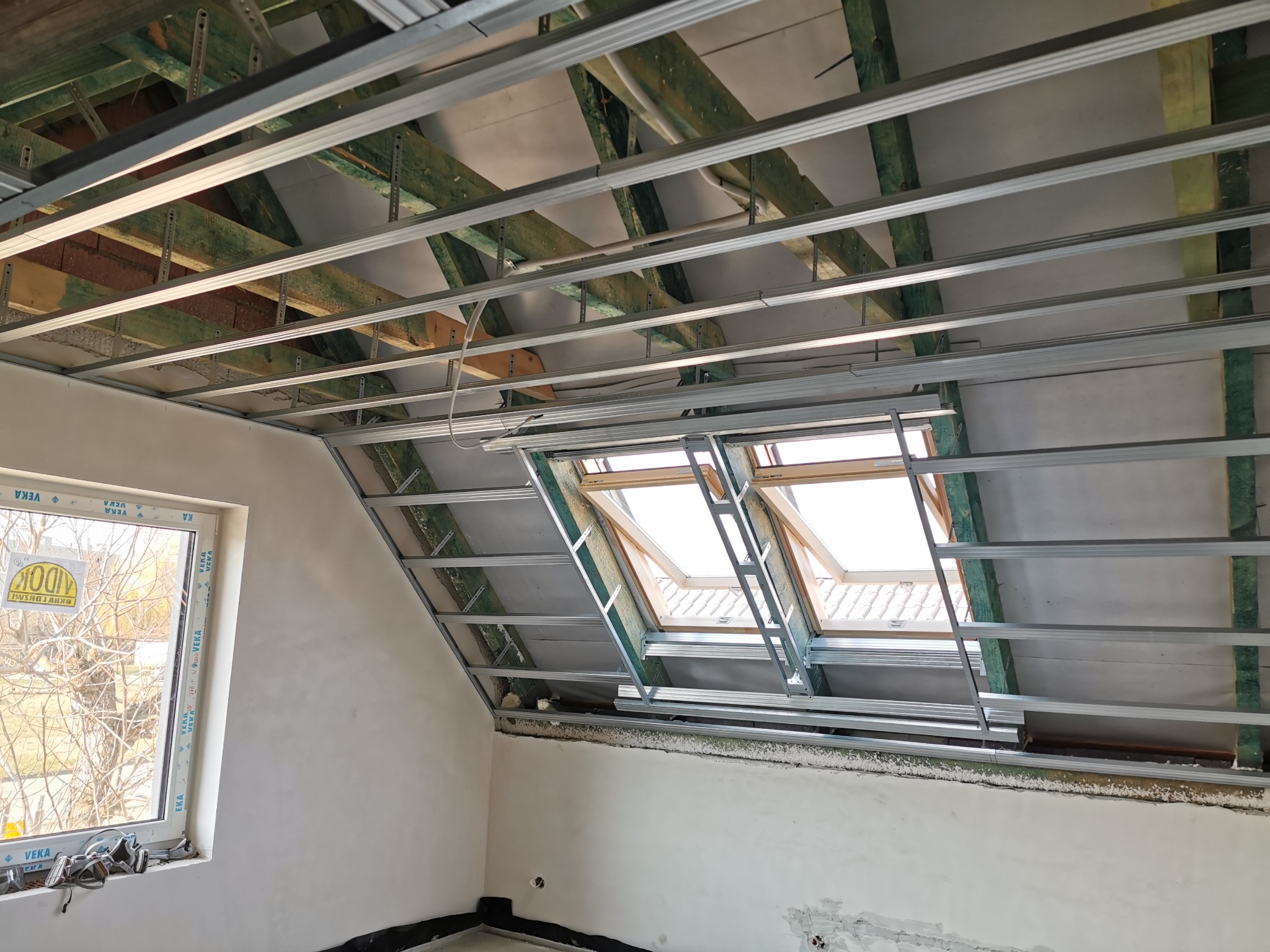 Montaż metalowej konstrukcji pod płyty gipsowo-kartonowe na poddaszu z oknem dachowym, widoczne drewniane elementy konstrukcyjne dachu i fragment ściany z oknem.