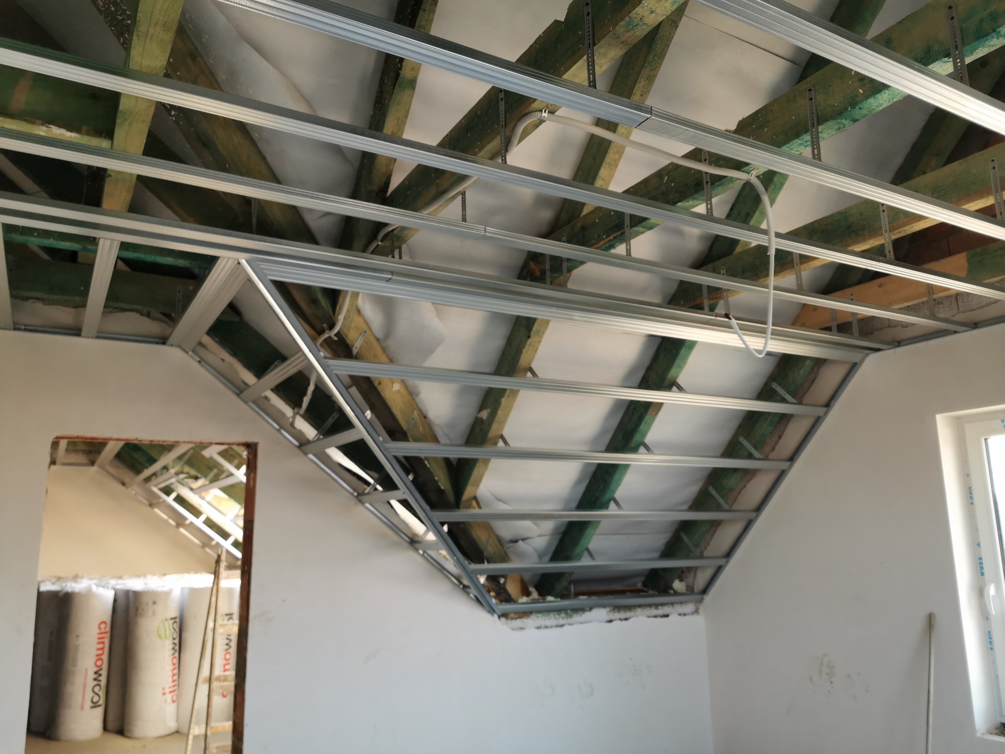 Konstrukcja metalowa pod płytę gipsową zamontowana na drewnianych belkach stropu w Rakszawie, widoczne rolki wełny mineralnej w sąsiednim pomieszczeniu.