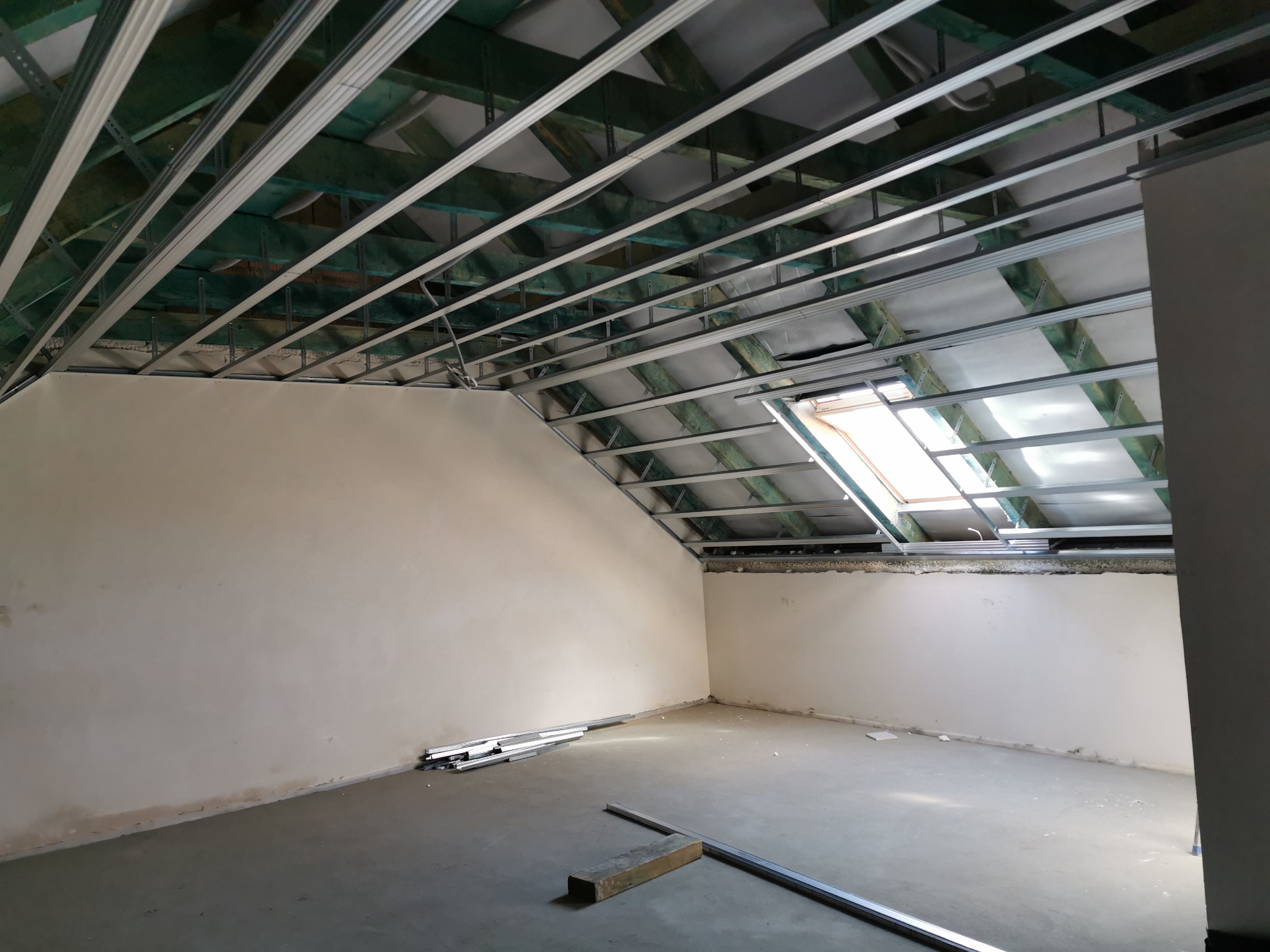 Poddasze w trakcie remontu: widoczne metalowe profile sufitowe na tle drewnianej konstrukcji dachu i okna dachowego, ściany częściowo wykończone, na podłodze leżą profile i fragment płyty.