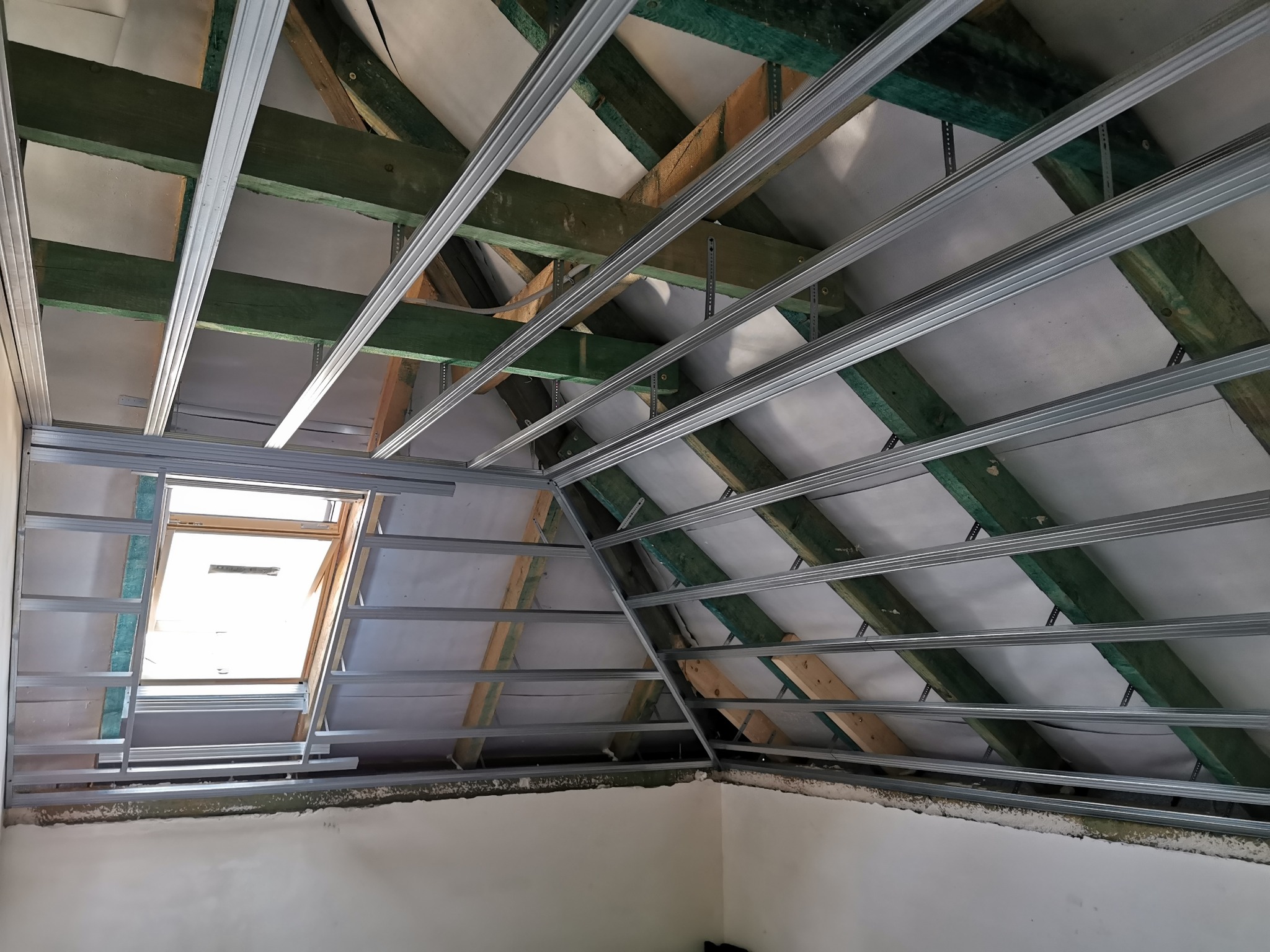 Konstrukcja metalowa pod płytę gipsowo-kartonową zamontowana na drewnianych belkach poddasza, widoczne okno dachowe i izolacja w tle.