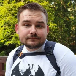 Mężczyzna z brodą w białej koszulce z grafiką i plecakiem na tle zieleni lasu. Portret w plenerze, naturalne światło.