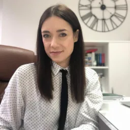 Portret kobiety w koszuli w kropki i krawacie, siedzącej przy biurku z zegarem na ścianie. Uśmiechnięta twarz, profesjonalny wygląd, biuro w tle.