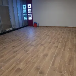 Wyłożona drewnopodobnymi płytkami podłoga w lokalu po remoncie, widoczne ślady po pracach budowlanych, resztki materiałów, w tle logo 'Big Star'.