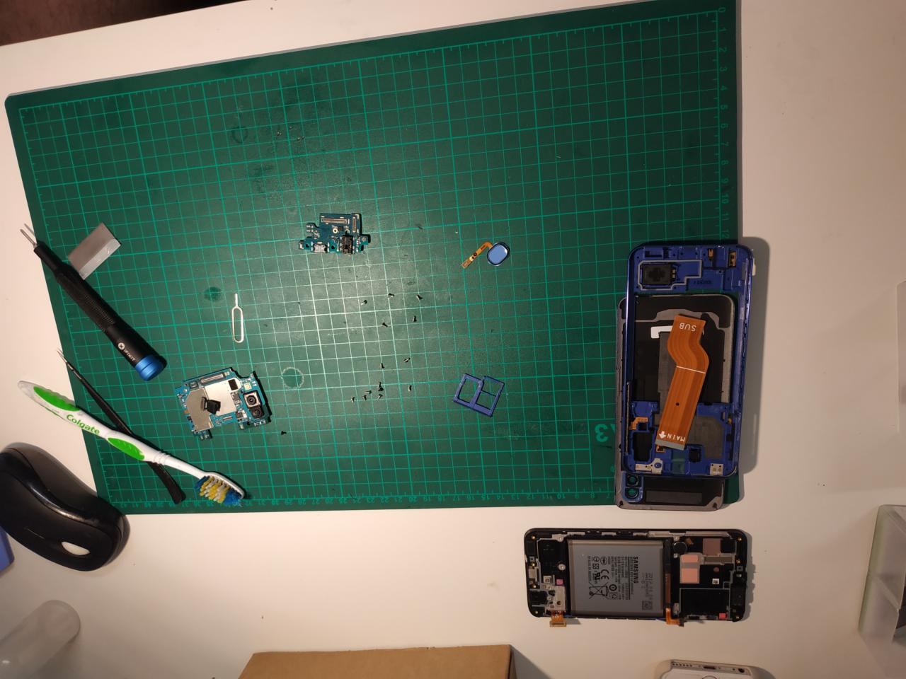 Rozłożony telefon Samsung na zielonej macie do cięcia, widoczne narzędzia: pęseta, śrubokręt i szczoteczka do zębów Colgate. Elementy telefonu porozrzucane obok obudów.
