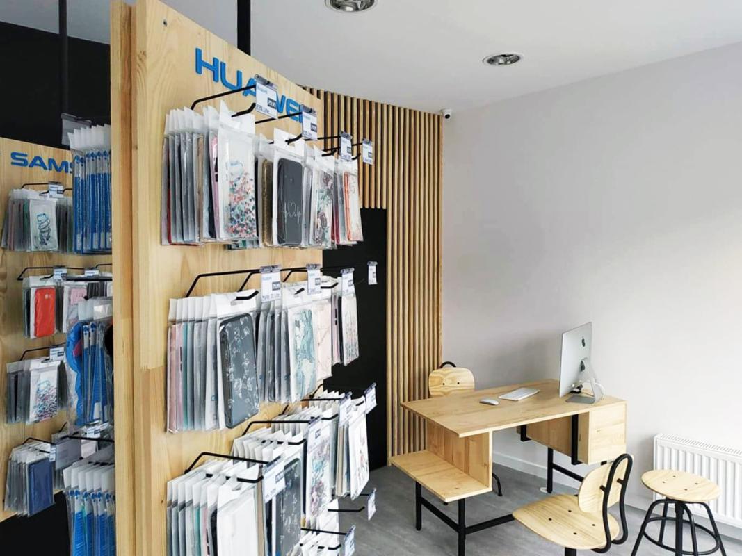 Wnętrze sklepu z akcesoriami do telefonów komórkowych: drewniane regały z etui na telefony marek Huawei i Samsung, proste biurko z komputerem i krzesłami.