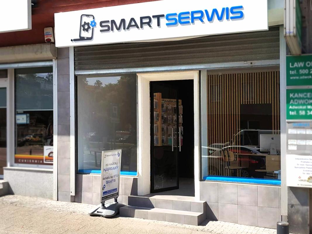Fasada serwisu telefonów SmartSerwis z logo przedstawiającym telefon z kołem zębatym, tablica reklamowa z napisem 'Do każdej naprawy dowolne etui gratis', widok na wnętrze z akcesoriami...