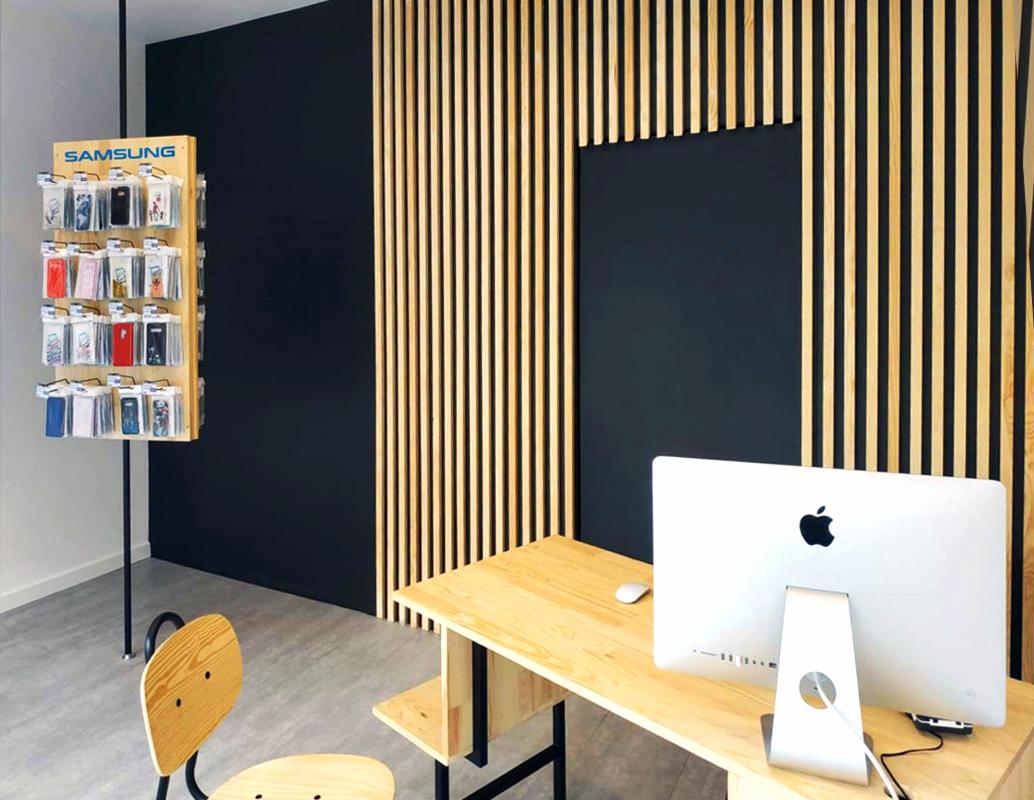 Wnętrze punktu usługowego: ekspozytor z etui na telefony Samsung, biurko z komputerem Apple iMac, czarne ściany z dekoracyjnymi drewnianymi listwami.