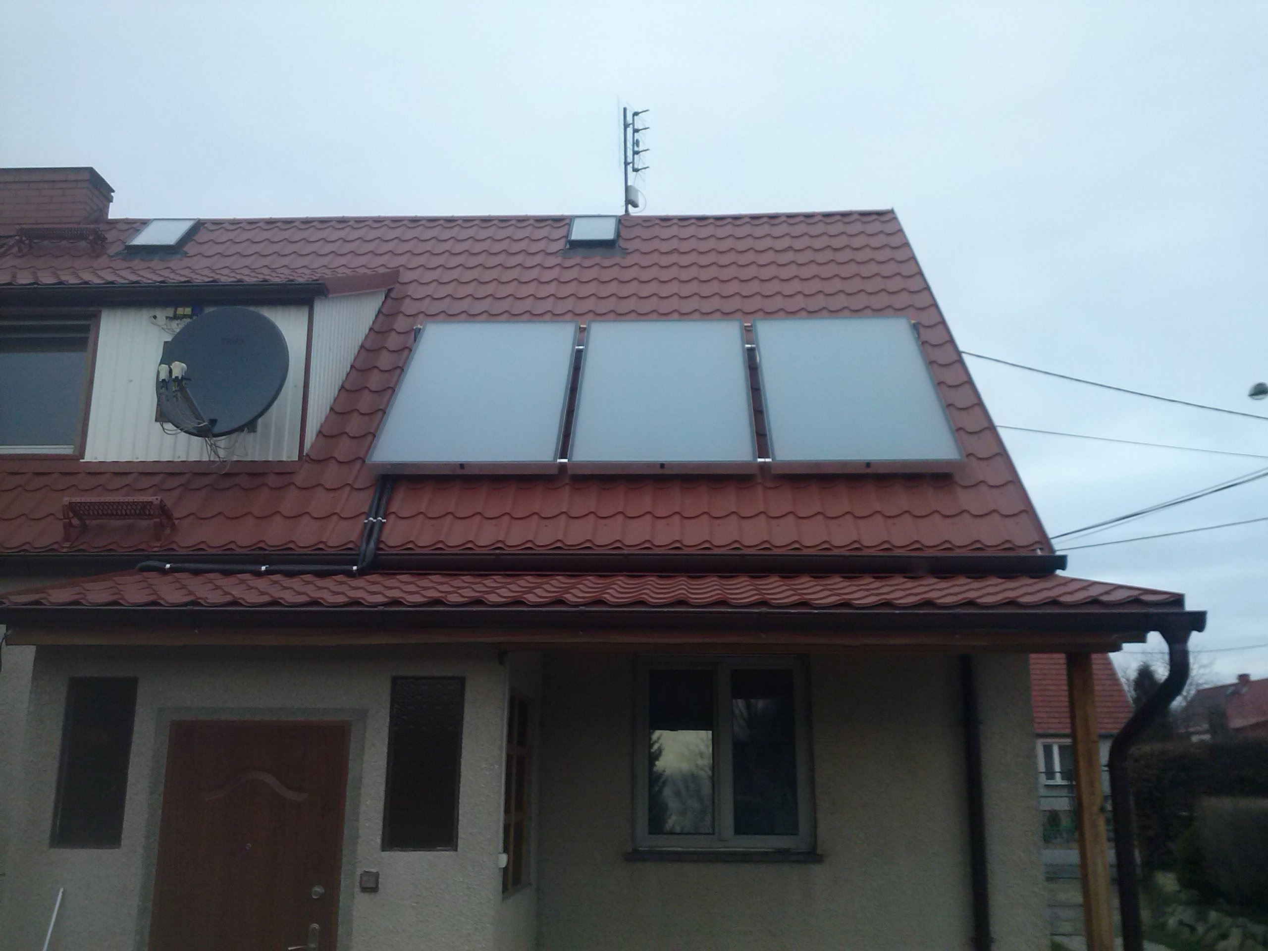 Dom jednorodzinny z trzema panelami solarnymi zamontowanymi na dachu pokrytym czerwoną blachodachówką, antena satelitarna na ścianie bocznej, pochmurne niebo w tle.