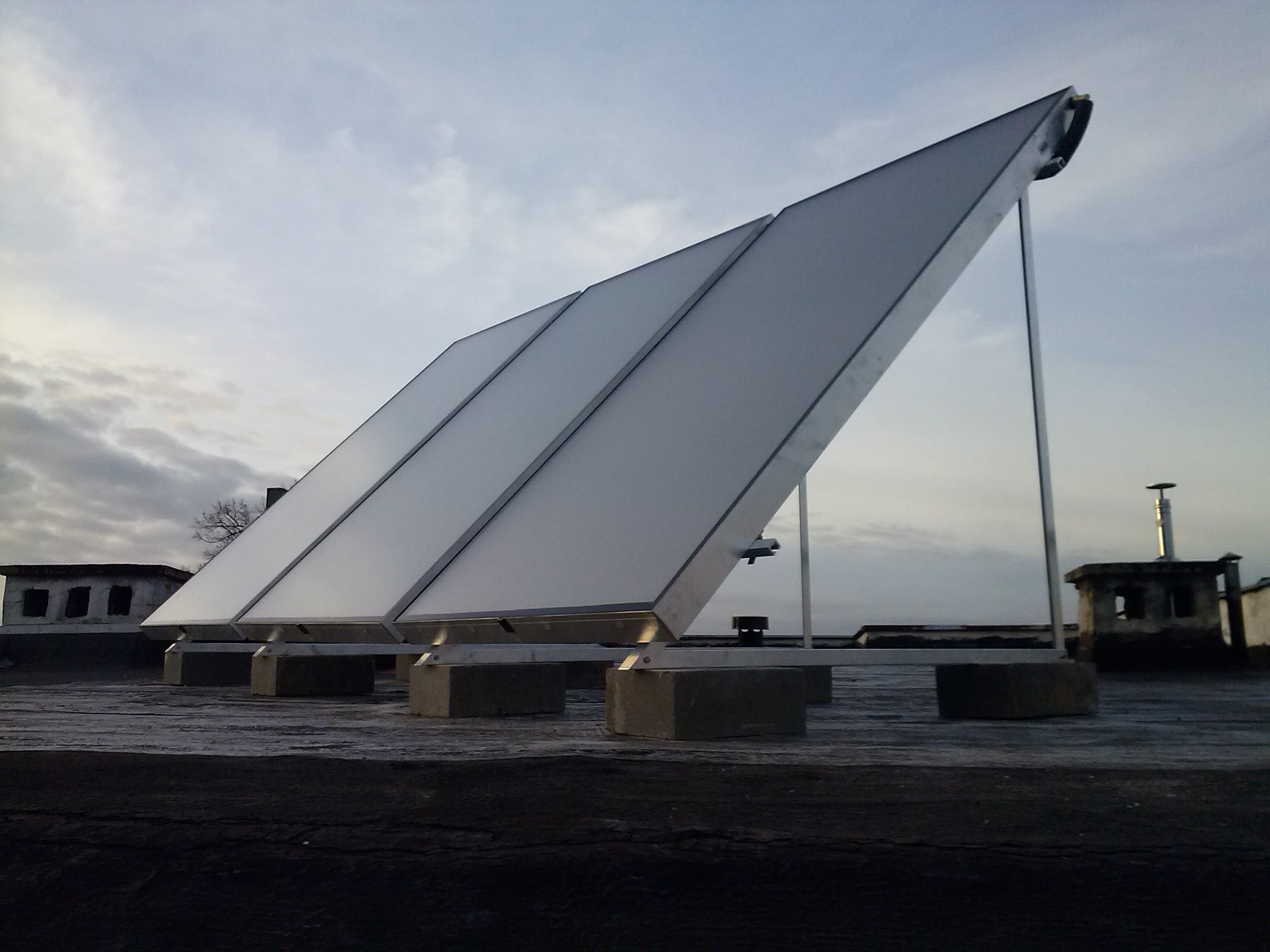 Trzy panele kolektorów słonecznych zamontowane na betonowych podstawach na płaskim dachu budynku, widoczne inne budynki w tle pod pochmurnym niebem.