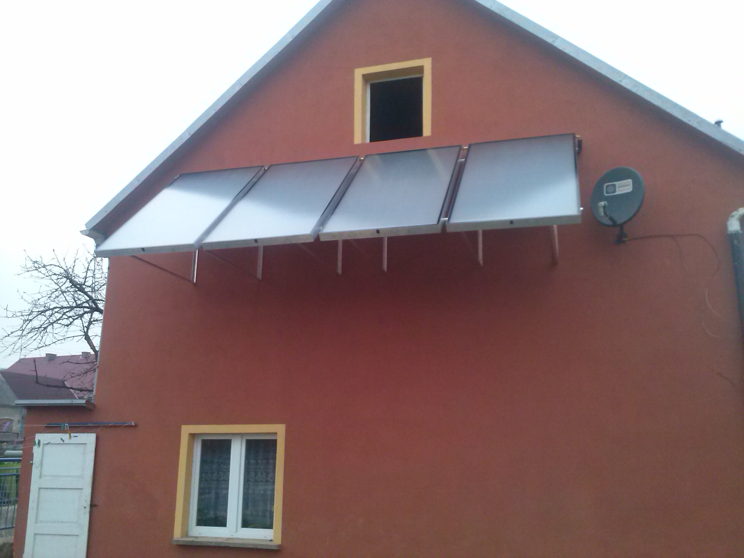 Trzy kolektory słoneczne zamontowane na ścianie domu z brązową elewacją, widoczna antena satelitarna i okna z żółtymi ramami.