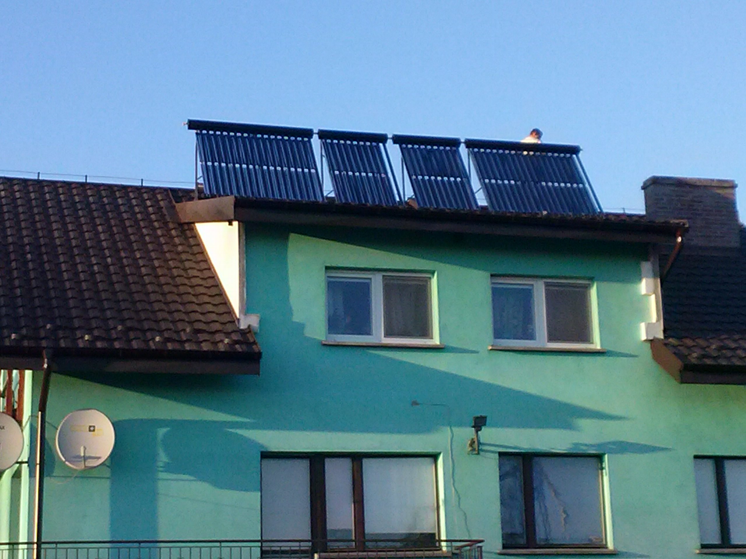Cztery kolektory słoneczne rurowe zamontowane na dachu dwupiętrowego, turkusowego domu z brązową dachówką, pod błękitnym niebem; widoczna osoba na dachu przy kolektorach.