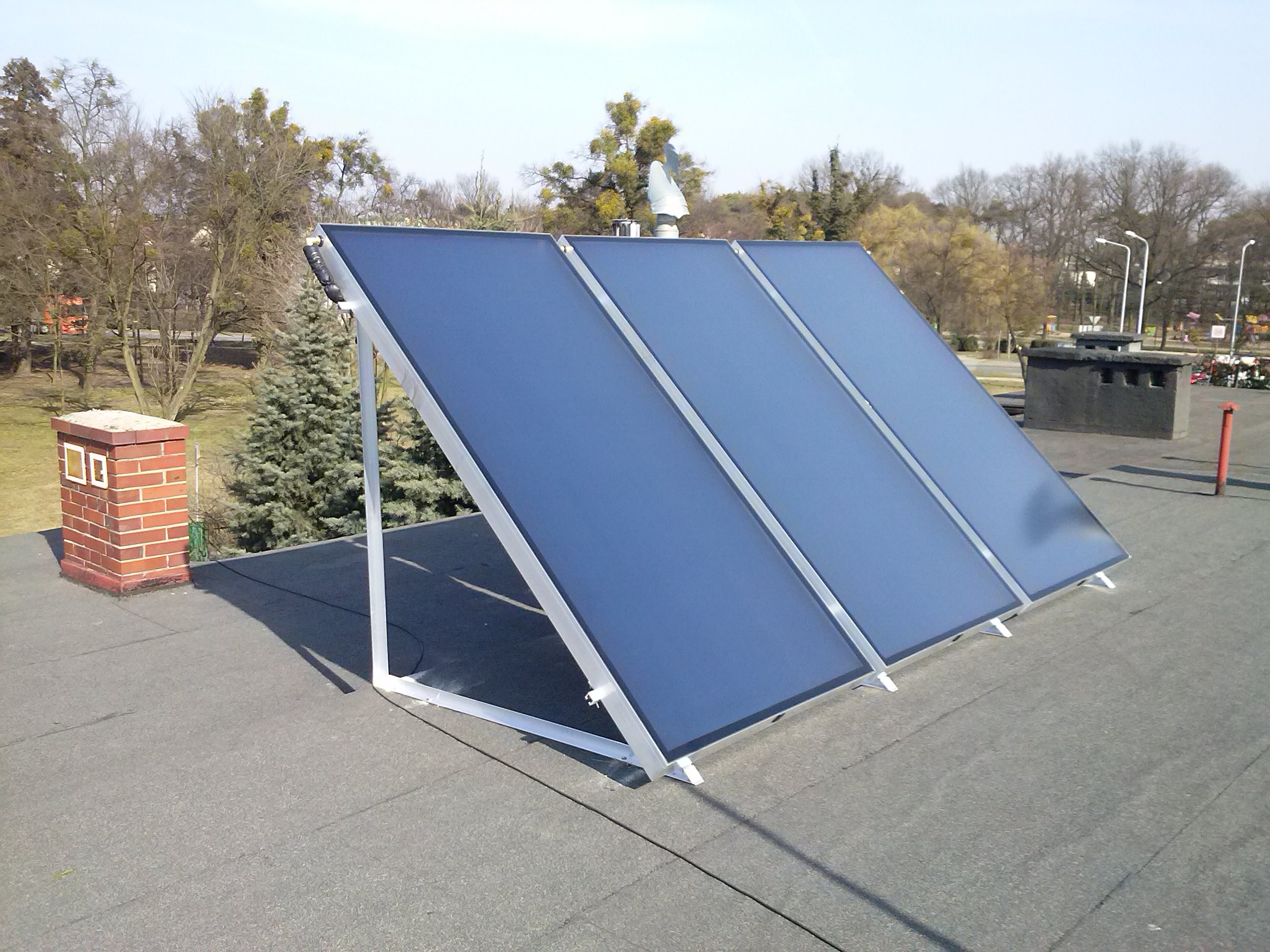 Trzy kolektory słoneczne zamontowane na płaskim dachu budynku, widoczny komin z cegły i drzewa w tle.