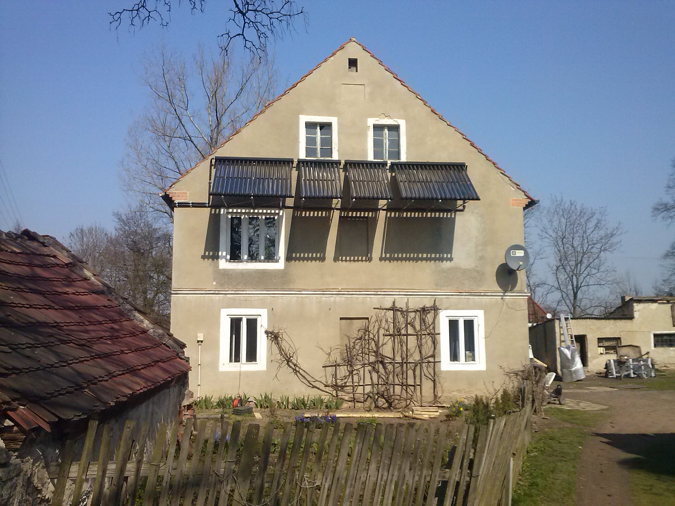 Dom jednorodzinny z zamontowanymi trzema panelami kolektorów słonecznych na elewacji, widoczne także antena satelitarna i drewniana konstrukcja pod pnącza.