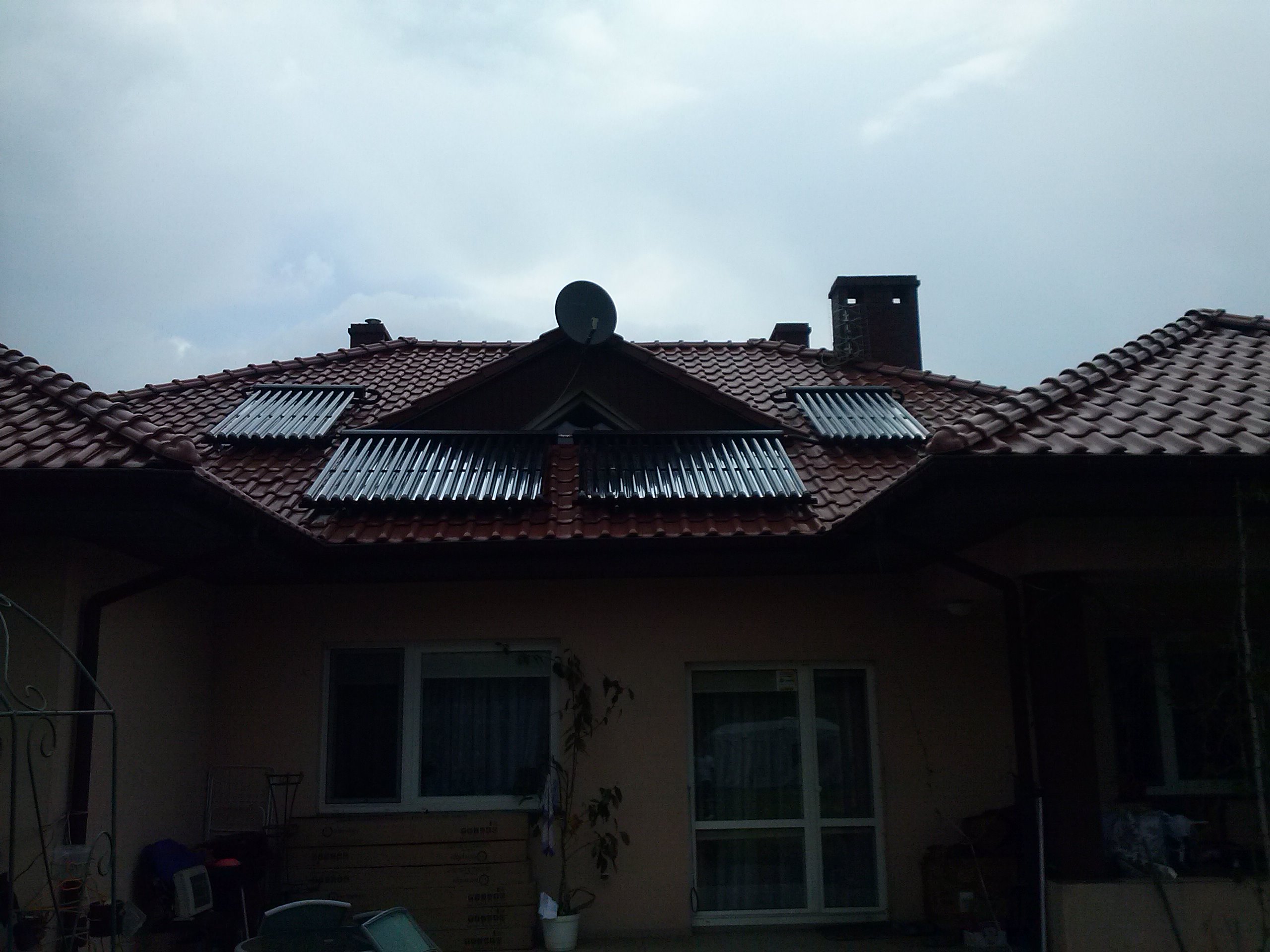Dom jednorodzinny z kolektorami słonecznymi na dachu z czerwonej dachówki, widoczna antena satelitarna i komin, pochmurne niebo.