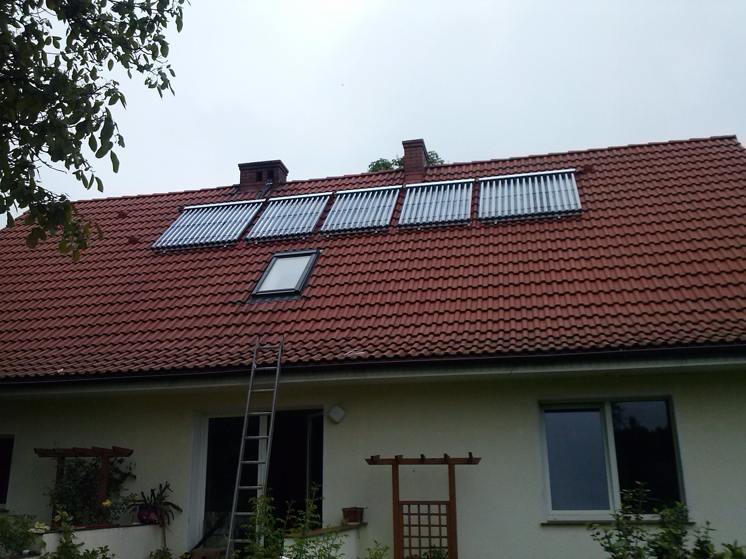 Czerwony dach domu jednorodzinnego z zamontowanymi trzema panelami kolektorów słonecznych, kominem i oknem dachowym; drabina oparta o ścianę budynku.